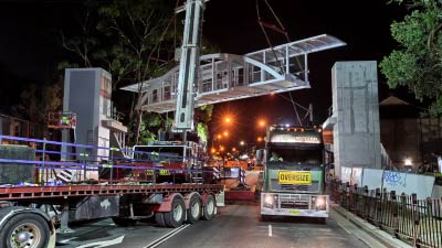 Cranes lift a large bridge over a highway for a walkway.