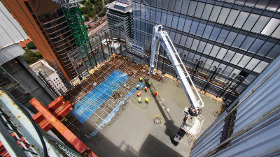Scaffolding-mounted camera view of workers pouring a concrete floor beside city buildings